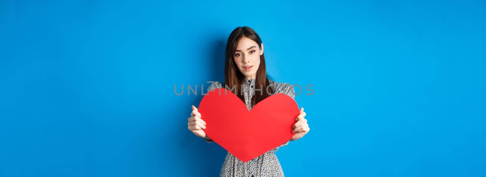 Valentines day and relationship concept. Tender young woman in dress stretch out hand and giving big red heart to you, making confession, standing romantic on blue background by Benzoix