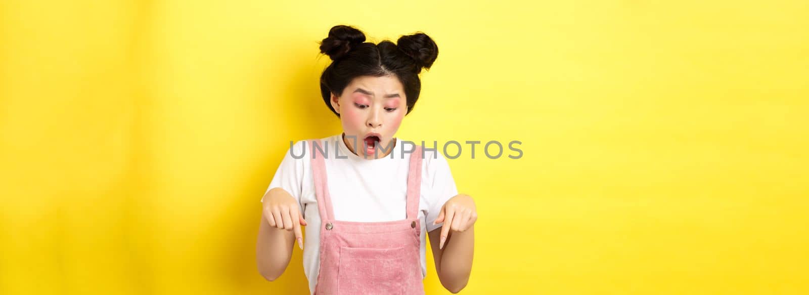 Excited teenage asian girl checking out advertisement, pointing fingers down and drop jaw amazed, showing promo offer, yellow background by Benzoix