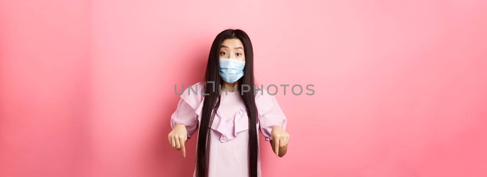 Healthy people and covid-19 pandemic concept. Excited asian girl in face mask pointing fingers down, showing advertisement with interest, standing against pink background.