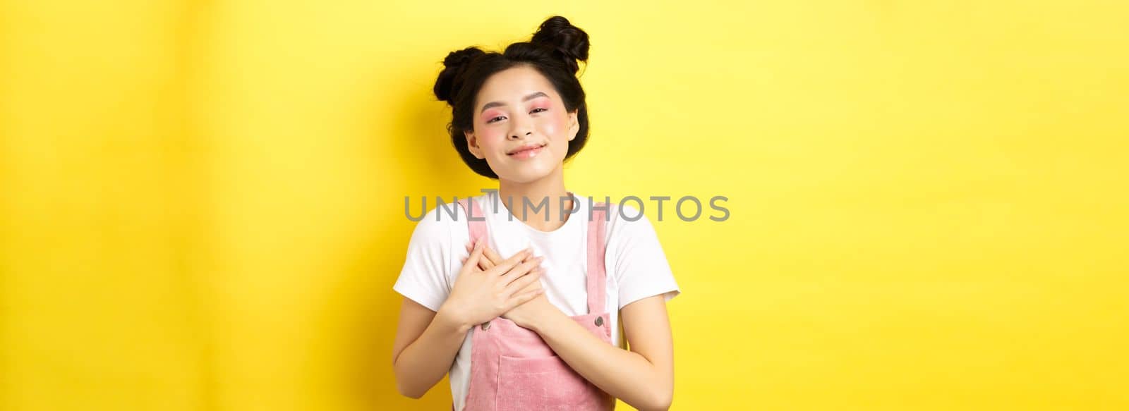 Beautiful teen asian girl in love, holding hands on heart and smiling touched, looking with affection, standing on yellow background by Benzoix