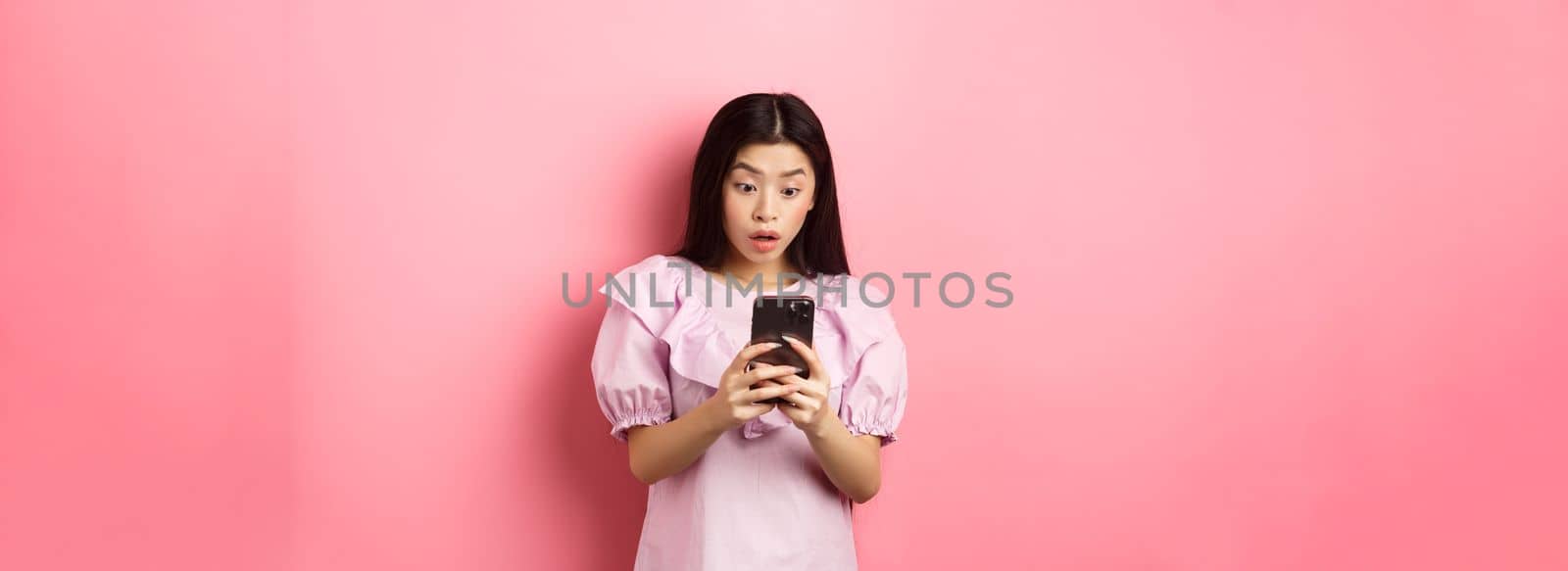 Online shopping. Asian teen girl look surprised at mobile phone screen, reading message with excited expression, standing against pink background by Benzoix