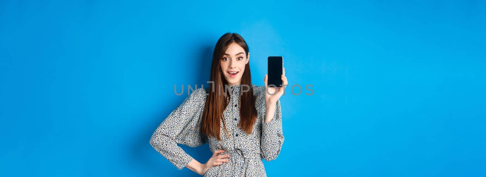 Amazed smiling woman showing empty smartphone screen, look impressed, recommending online promo, blue background by Benzoix