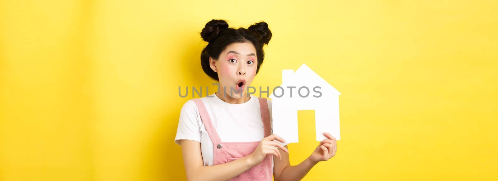 Real estate and family concept. Excited asian teen girl showing paper house cutout and gasping amazed, standing on yellow background by Benzoix