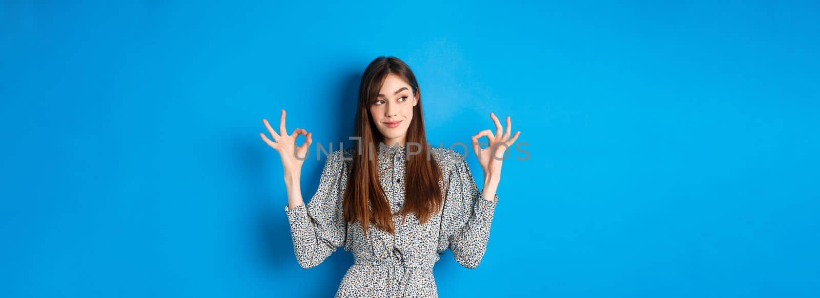 Not bad. Pleased smiling woman with long hair, wearing dress, showing okay signs and looking satisfied, praising good job, looking left at logo, blue background.