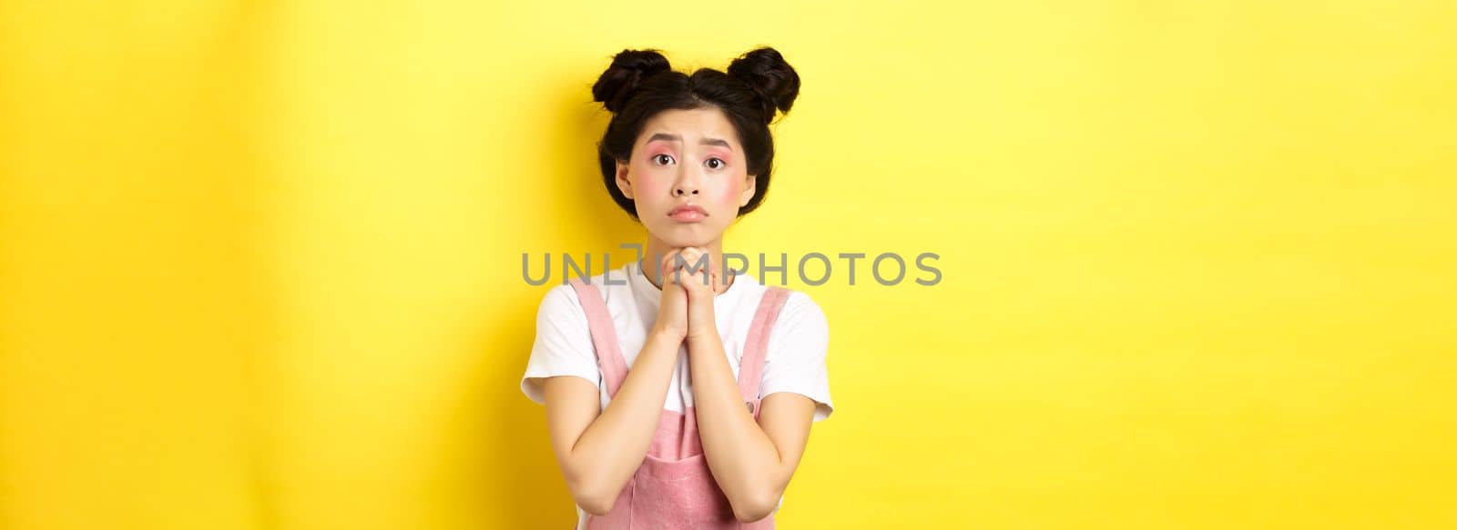 Hopeful asian stylish girl holding hands in begging sign, waiting with hope, asking for favour or help, standing on yellow background by Benzoix