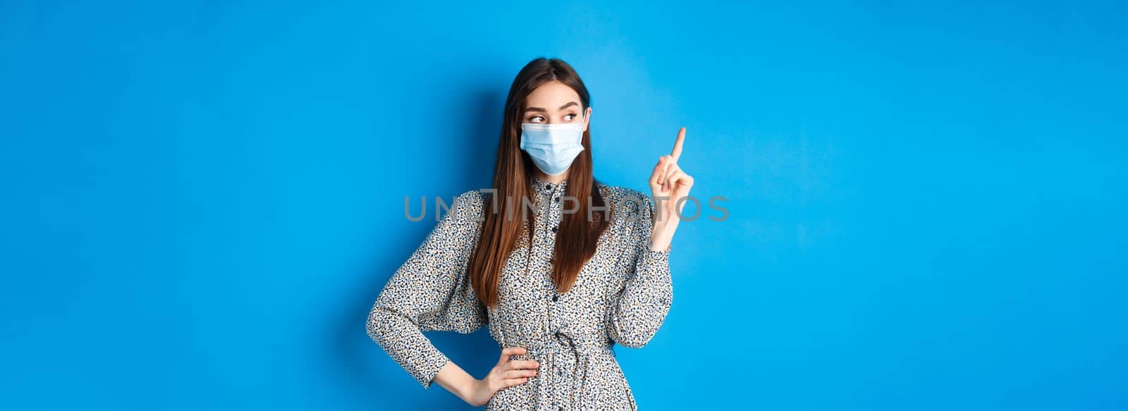 Healthy people and covid-19 pandemic concept. Curious girl in medical mask pointing at upper left corner, standing on blue background.