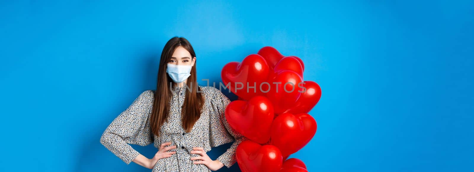 Pandemic and Valentines day. Cheerful smiling girl in medical mask, standing near romantic heart balloons and looking at camera, wearing dress, blue background.