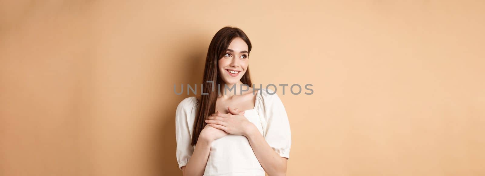 Romantic young girl in dress holding hands on heart, smiling and looking at empty space thankful, feeling gratitude, standing on beige background.