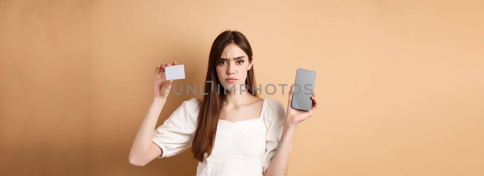 Sad and disappointed girl showing empty smartphone screen and plastic credit card, complaining and frowning, standing on beige background.