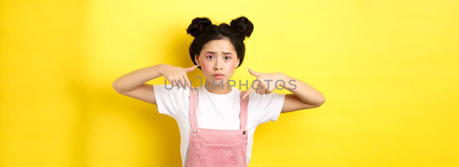 Worried and confused asian girl with glamour makeup, pointing fingers down and look sad, standing in summer clothes against pink background by Benzoix