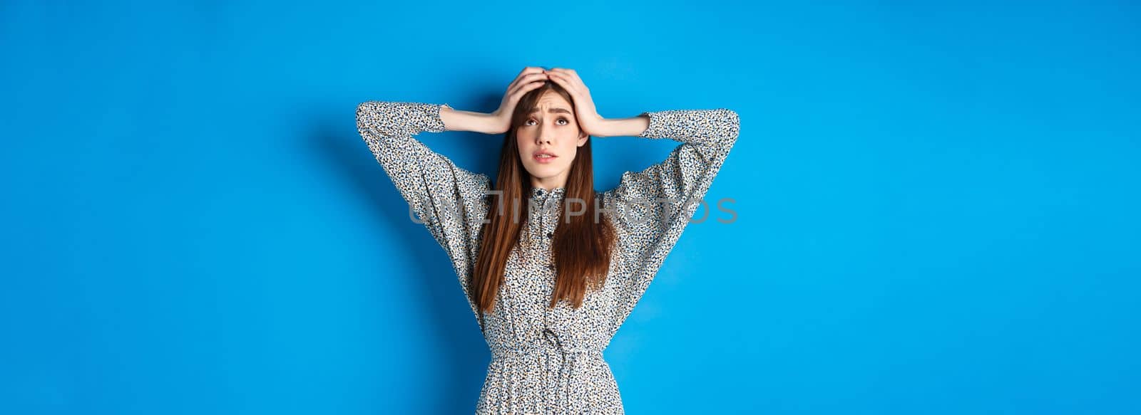 Troubled young woman with long hair, holding hands on head in panic, looking up nervous, having problem, standing on blue background.