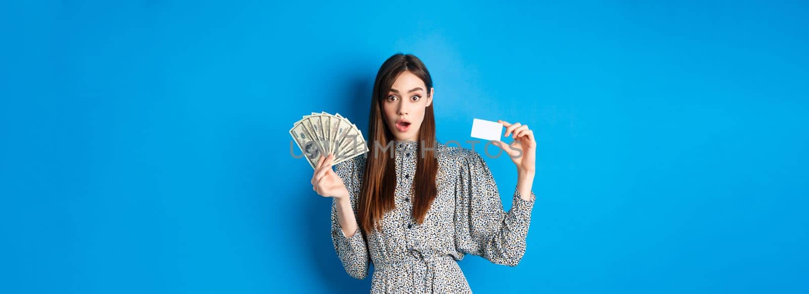 Excited lucky girl showing plastic credit card and money dollar, gasping amazed, buying something expensive, standing on blue background by Benzoix