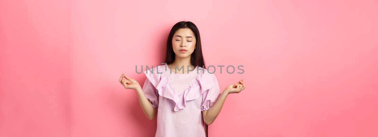 Stay calm. Peaceful asian woman meditating, hold hans in zen gseture and close eyes, practice yoga, standing patient on pink background by Benzoix