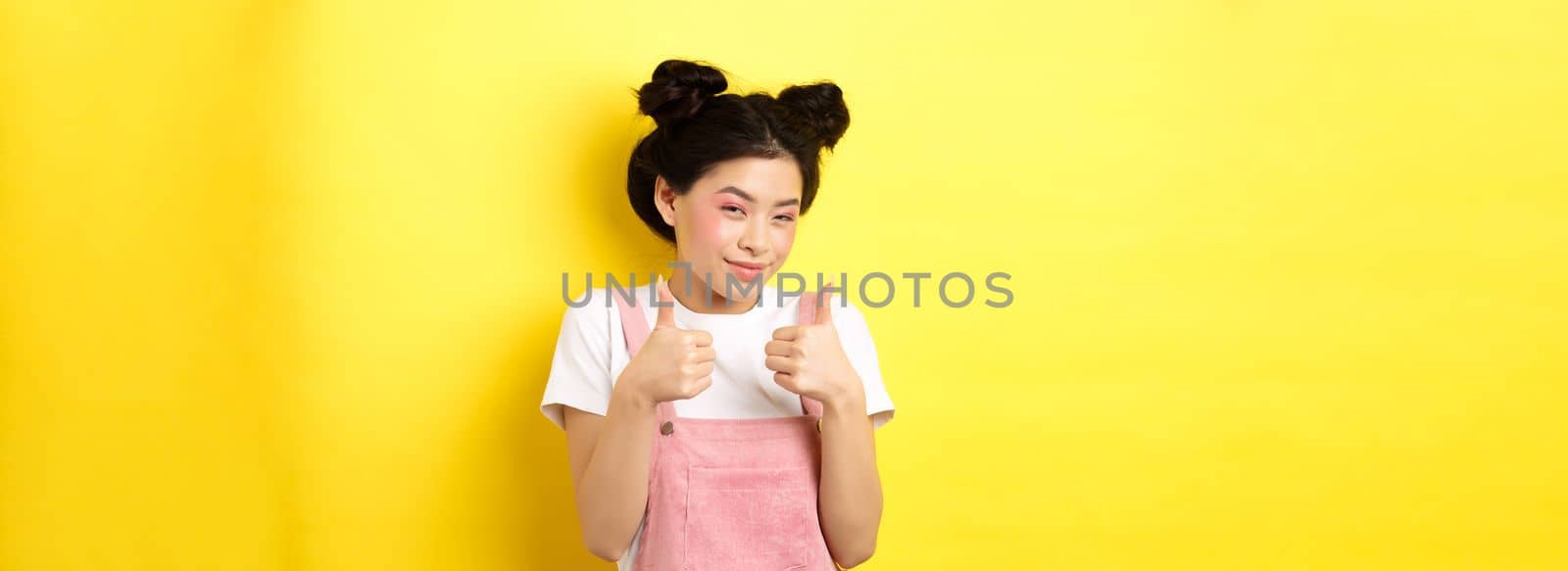 Cute teen asian girl with beauty makeup, showing thumbs up and smiling, praising and motivating you, making compliment, standing pleased on yellow background by Benzoix