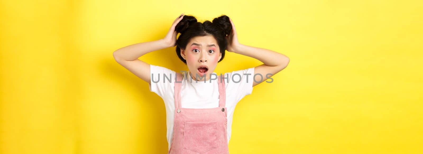 Shocked asian girl gasping worried, checking her hairstyle, standing anxious on yellow background.