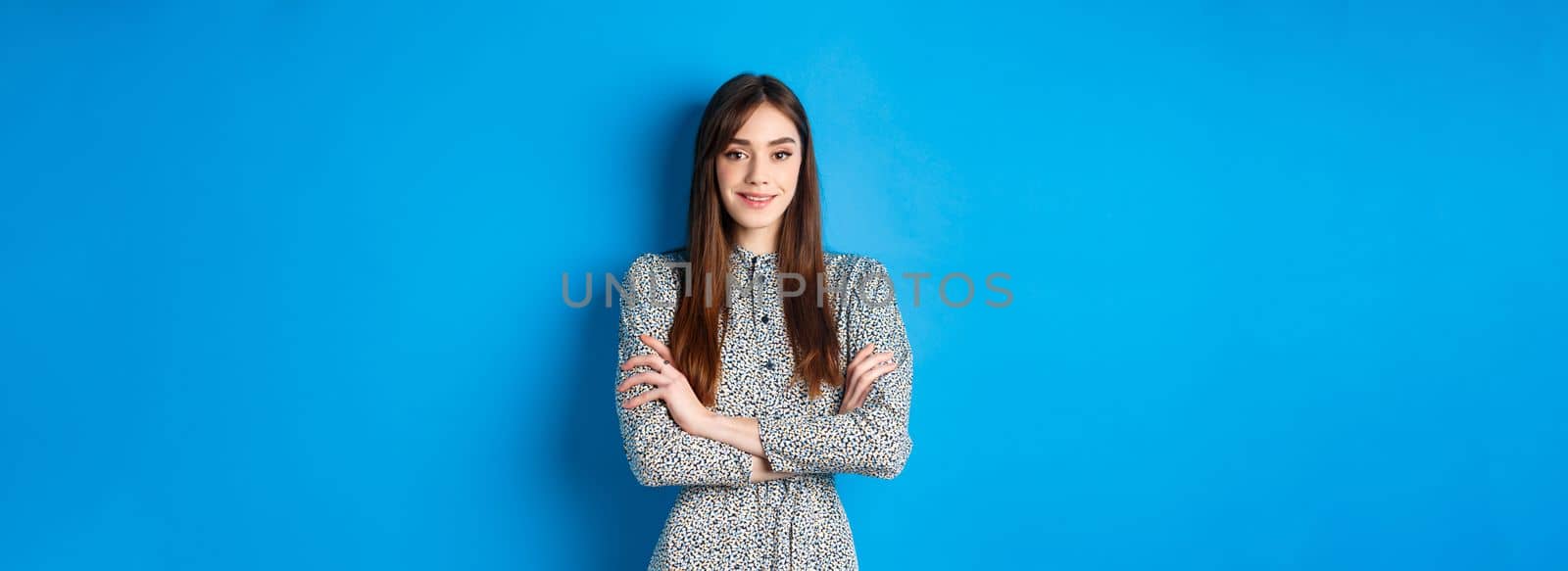 Elegant caucasian girl in dress cross arms on chest, standing on blue background.