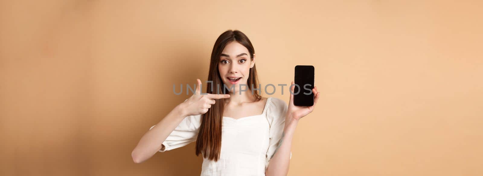 Cheerful woman pointing finger at empty phone screen, looking excited, standing on beige background.
