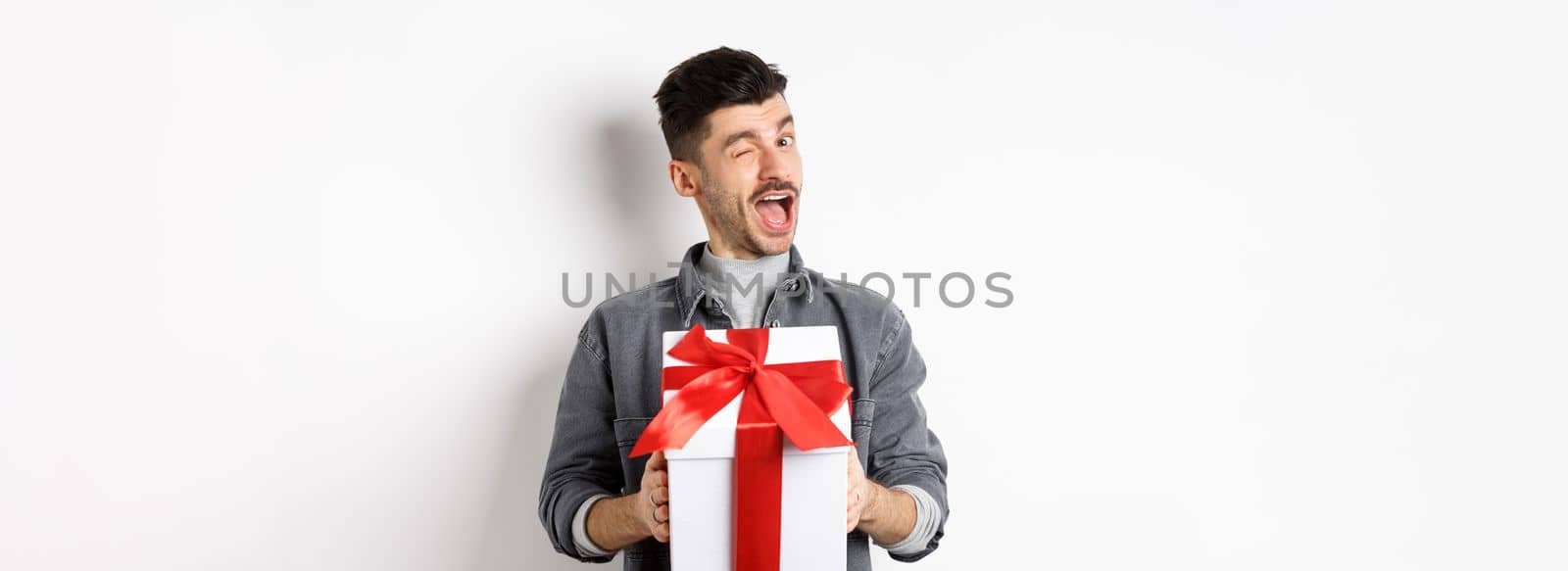 Funny young man holding valentines gift and winking at camera, making romantic surprise to lover, standing on white background by Benzoix