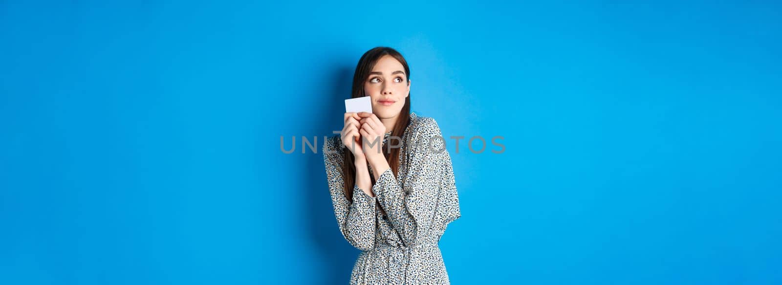 Romantic cute girl in dress dreaming of shopping, holding credit card and looking at logo with tender smile, standing on blue background.