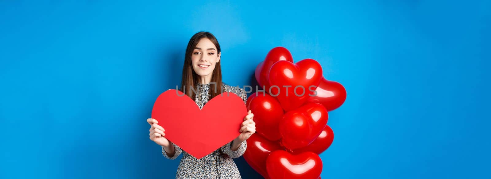 Valentines day. Lovely young woman in dress celebrating lovers holiday, showing valentine card and smiling, standing near red heart balloons on blue background.