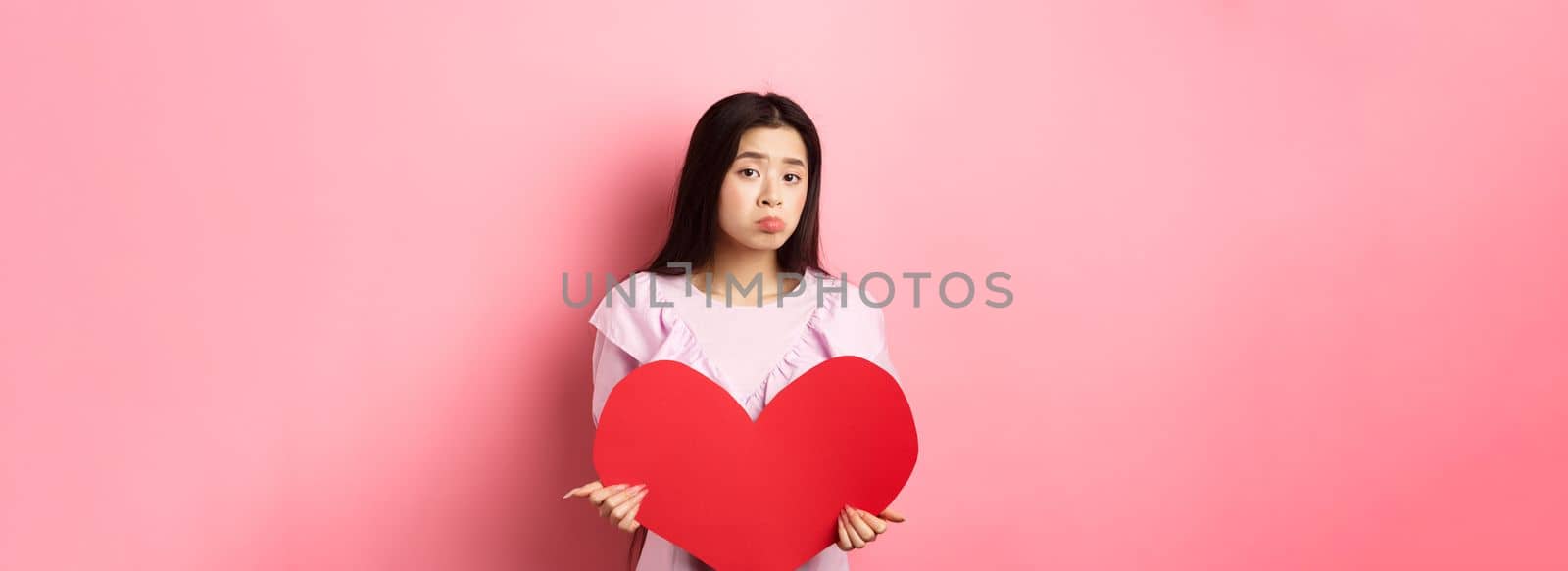 Valentines concept. Single teenage asian girl wants to fall in love, looking sad and lonely at camera, sulking distressed on lovers day, holding big red heart cutout, pink background by Benzoix