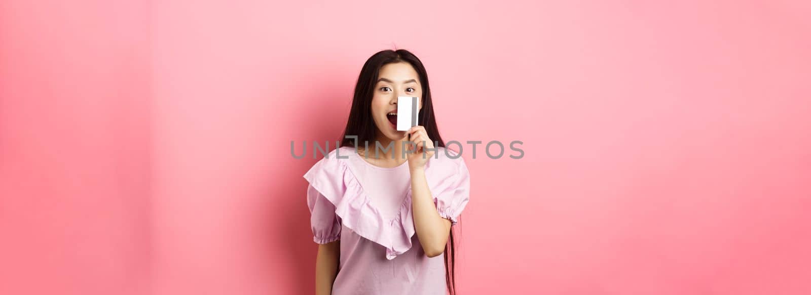 Excited asian girl showing plastic credit card and smiling, going on shopping, standing against pink background.