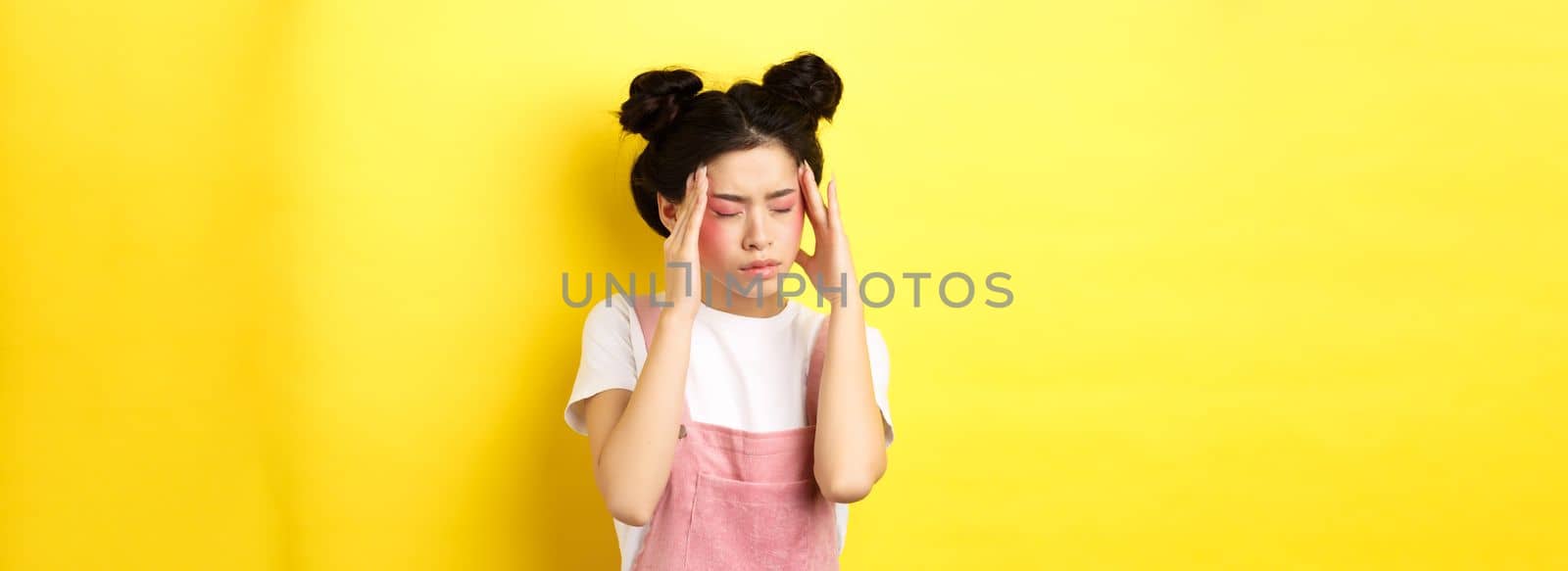 Image of stylish asian woman with bright makeup, having headache, touching head and looking tired, have painful migraine, yellow background.