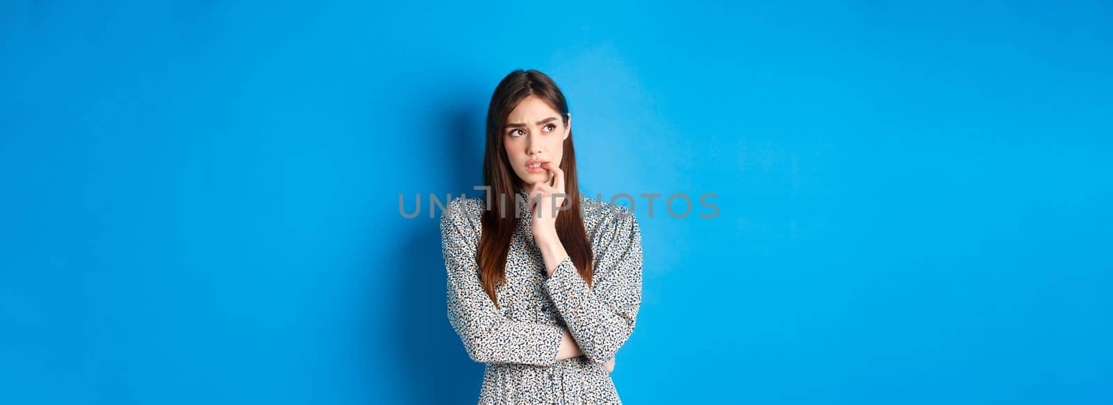Troubled pensive girl looking aside at logo, biting fingernail and thinking, standing on blue background in long dress.