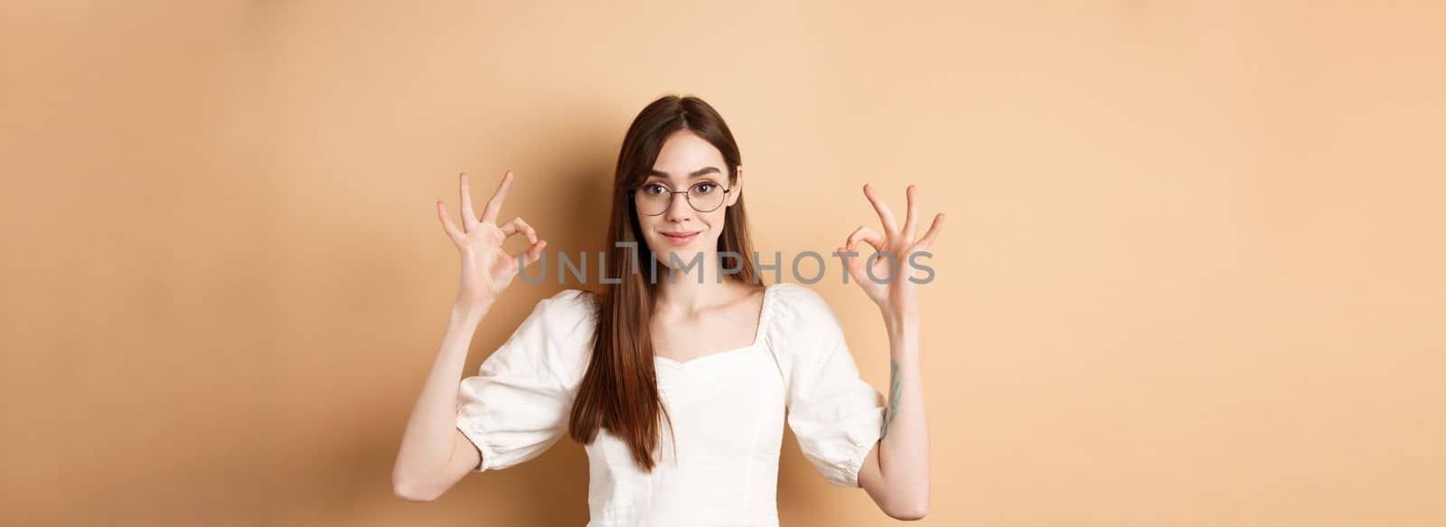 Satisfied female model in glasses show okay sign, looking pleased, agree and approve, standing on beige background by Benzoix