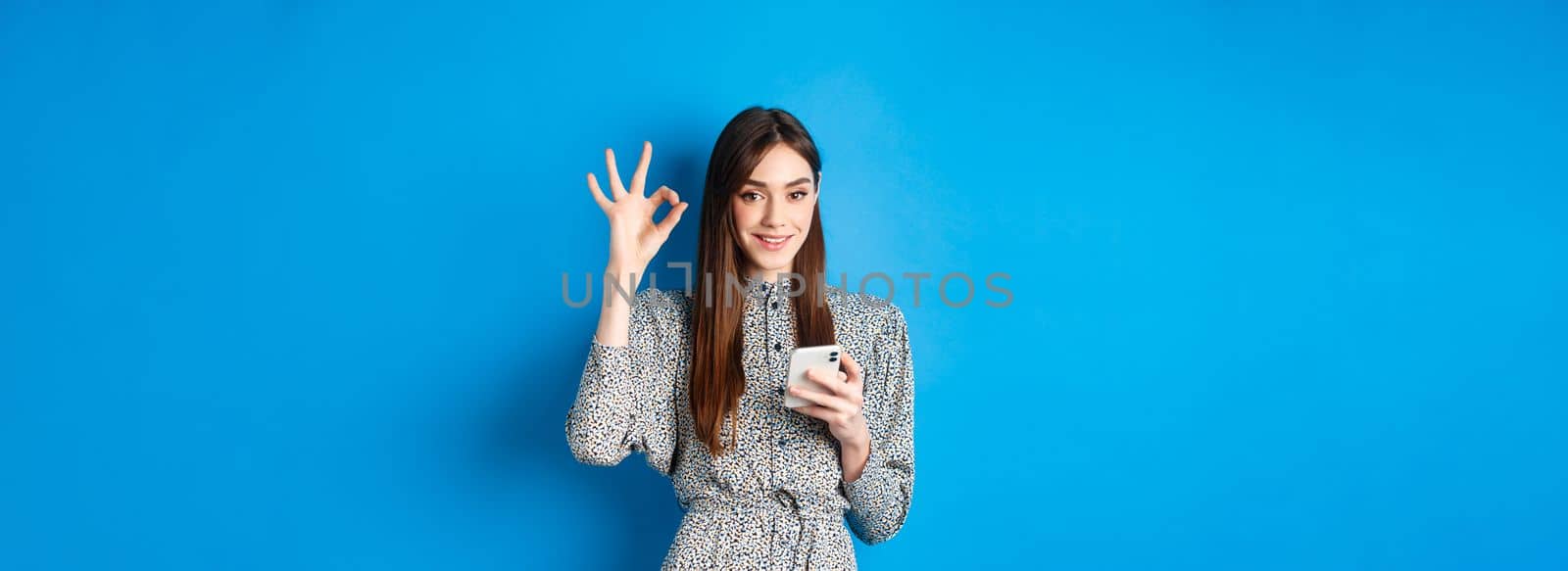 Beautiful young woman in dress showing okay sign and using smartphone, smiling at camera, standing on blue background.