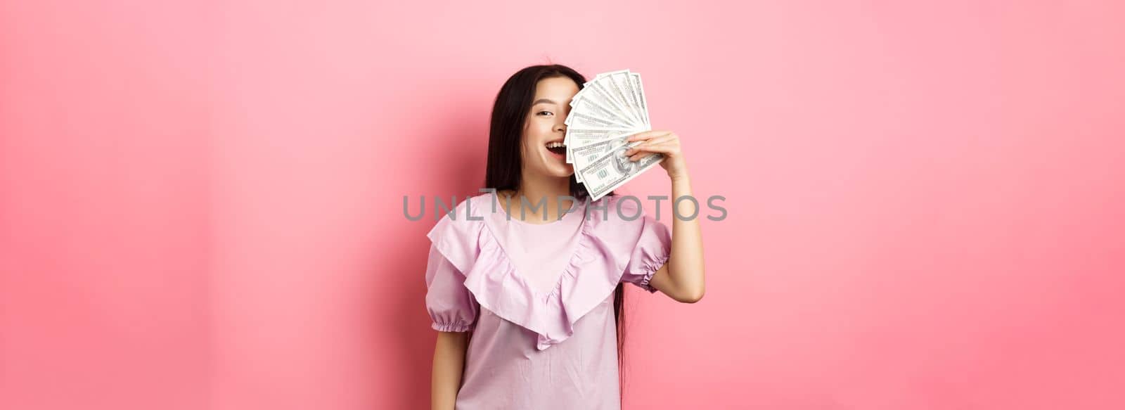 Happy rich asian woman showing money and smiling, shopping with cash, standing in dress against pink background.