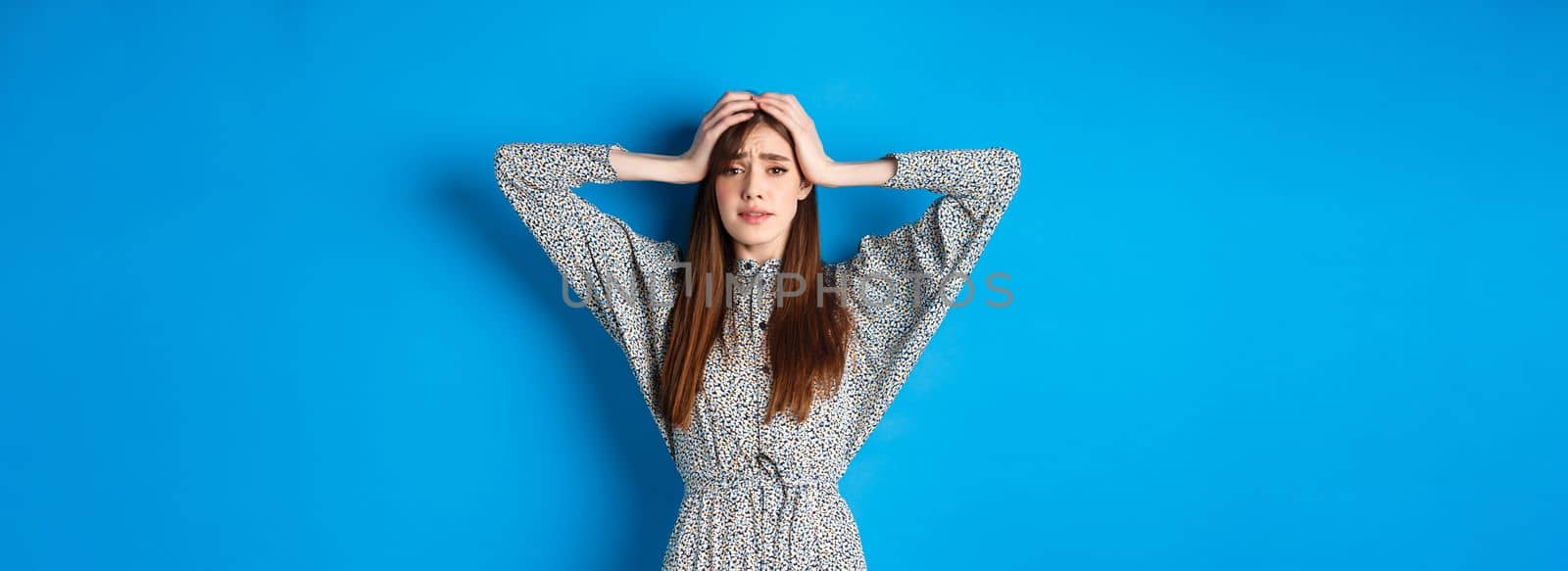 Frustrated adult woman with natural hair holding hands on head and panicking, looking alarmed and scared, standing in dress against blue background by Benzoix