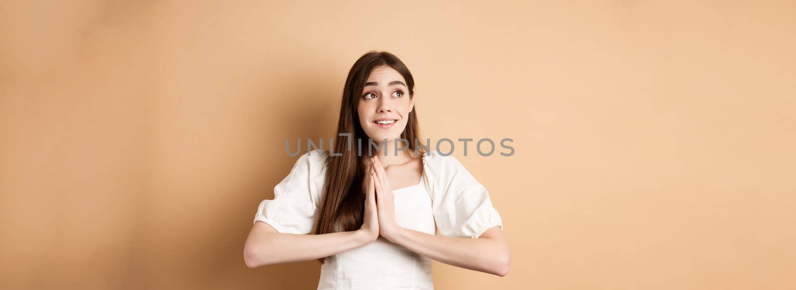 Hopeful young girl begging god and looking dreamy at upper left corner, praying or making wish, standing on beige background.