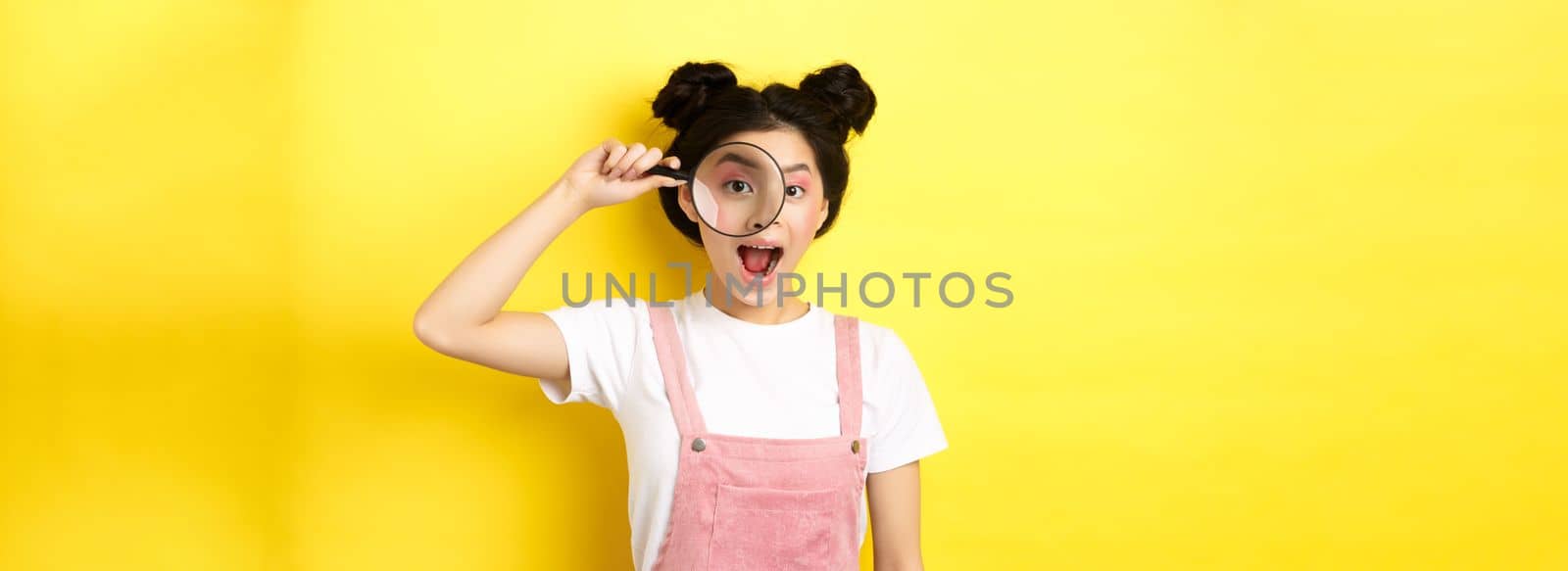 Young asian girl with bright makeup look through magnifying glass, smiling excited, seeing interesting promo, yellow background.