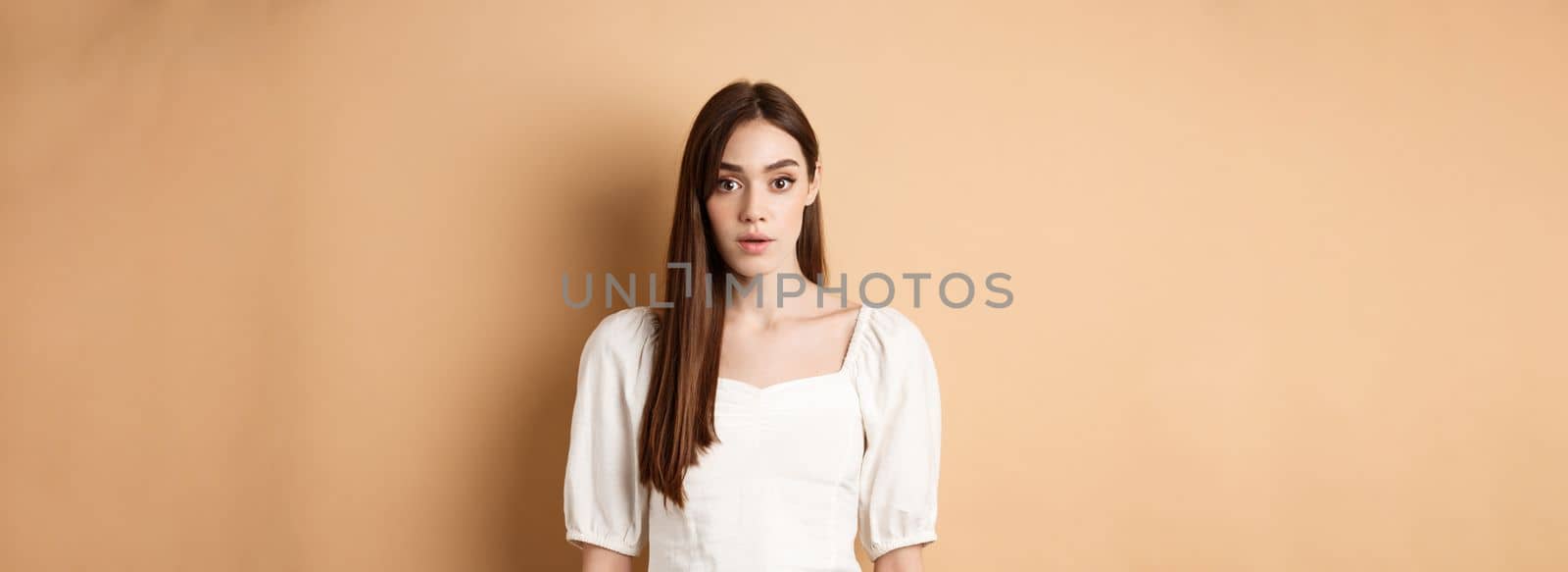 Shocked and startled young woman freeze in awe, open mouth and look at camera amazed, standing in dress on beige background.