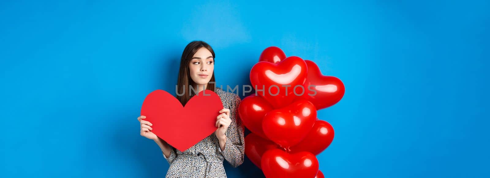 Valentines day. Romantic girl in dress standing near balloons and holding big red heart cutout, dream of something, standing on blue background by Benzoix