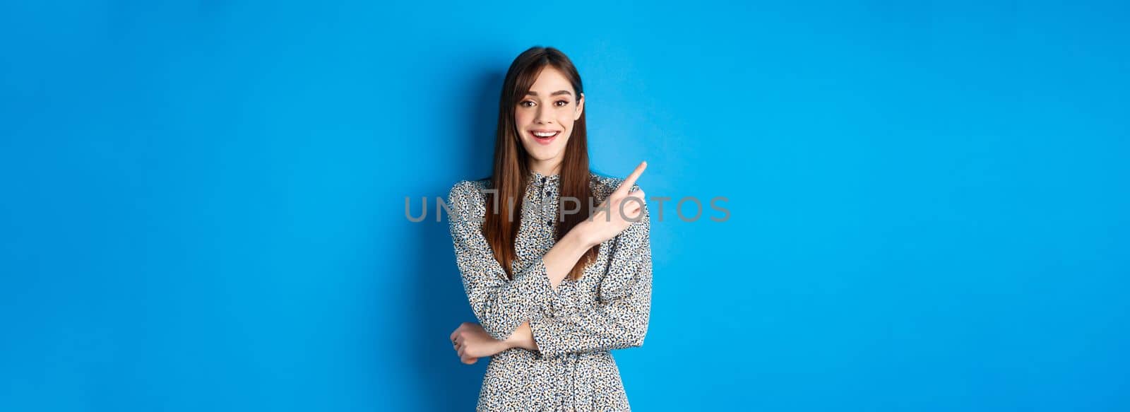 Cheerful young woman in long dress smiling, pointing finger at upper left corner, advertising product on blue background by Benzoix