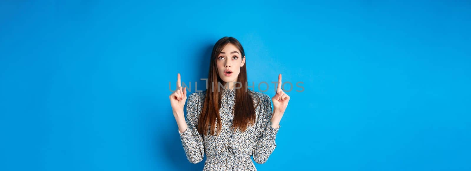 Surprised romantic girl in dress pointing, looking up with opened mouth and fascinated gaze, standing on blue background.