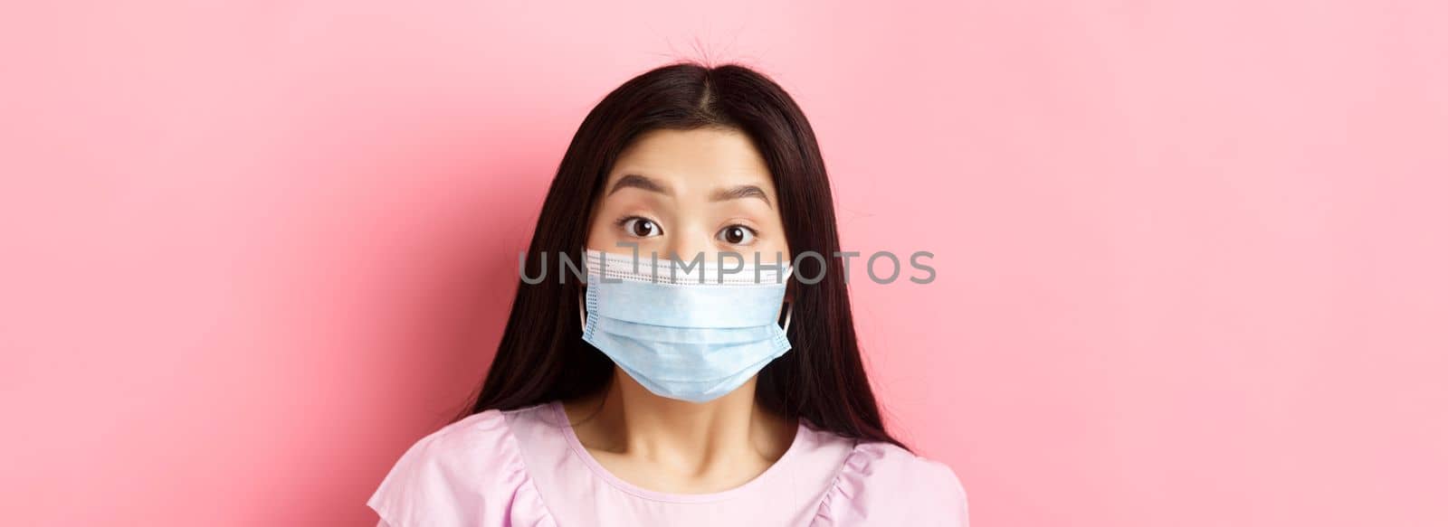 Covid-19 and healthy people concept. Close-up portrait of surprised asian girl in medical mask looking excited at camera, standing against pink background.
