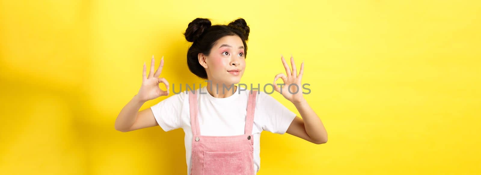 Romantic and beautiful asian girl looking at logo left, showing okay signs in approval, standing on yellow background.