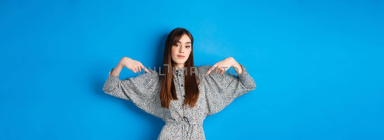 Confident pretty girl in dress pointing fingers at center logo, showing banner and looking self-assured, standing on blue background by Benzoix