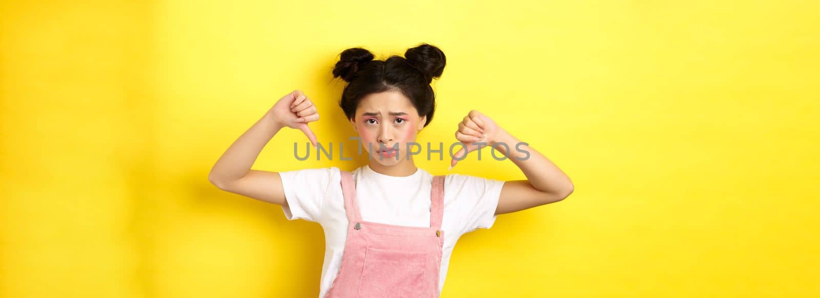 Summer lifestyle concept. Sad and gloomy asian woman with makeup, pointing fingers down and frowning upset, disappointed with unfair situation, yellow background.