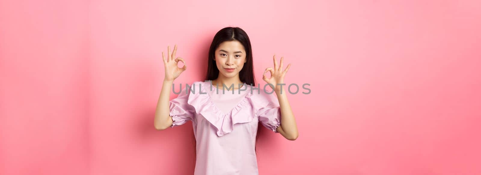 Smiling asian woman showing okay signs and looking confident, assure all good, praise good work, nice choice gesture, standing on pink background.