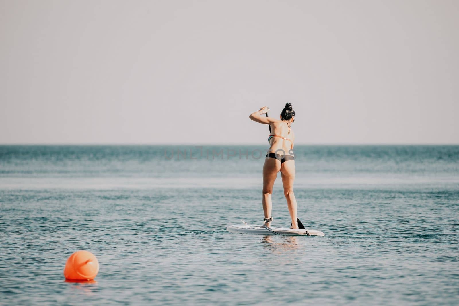 Sea woman sup. Silhouette of happy middle aged woman in rainbow bikini, surfing on SUP board, confident paddling through water surface. Idyllic sunset. Active lifestyle at sea or river