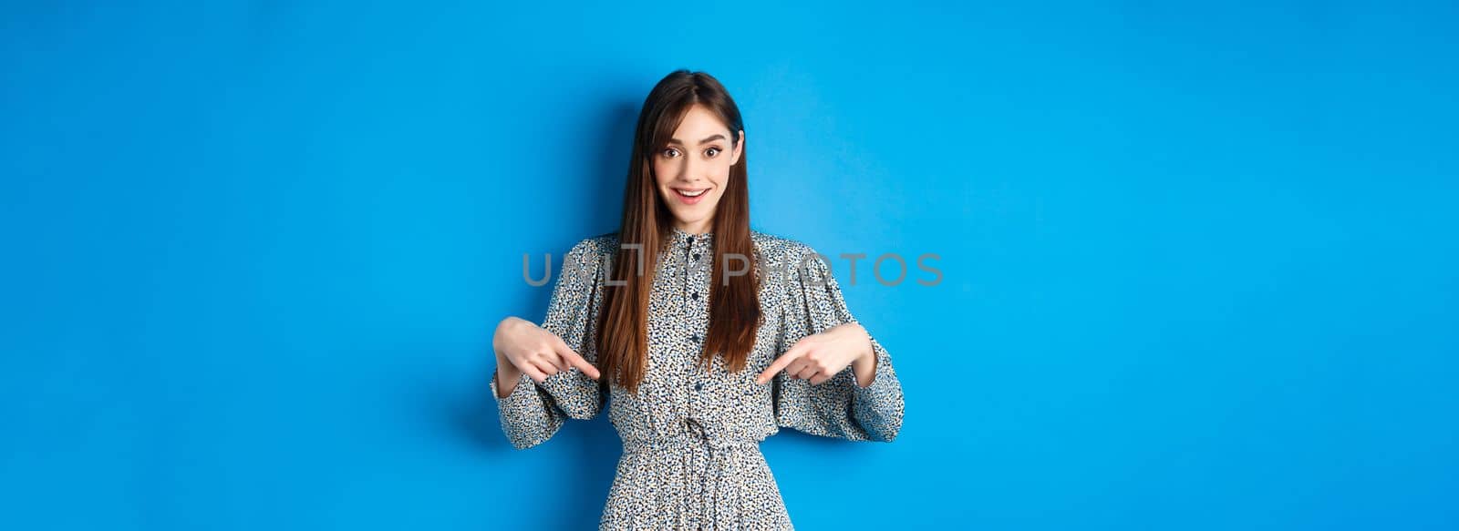 Excited caucasian woman in dress pointing fingers down at logo, showing fantastic news and smiling, standing on blue background.