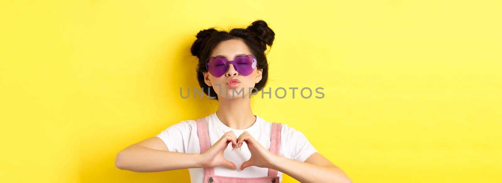 Summer and fashion concept. Cute glam girl with sunglasses and hairbuns, showing heart sign and pucker lips for kiss, I love you gesture, yellow background by Benzoix