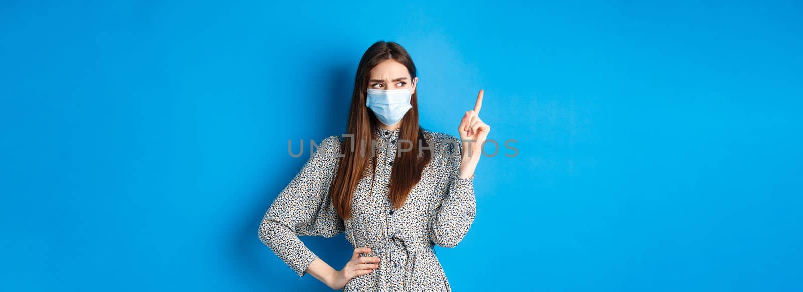 People, covid and quarantine concept. Suspicious frowning girl in medical mask, pointing and looking aside with hesitant face, standing on blue background by Benzoix