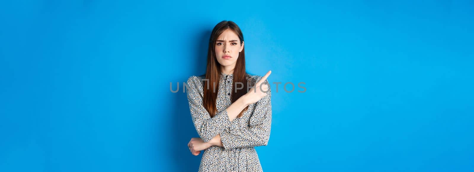 Disappointed frowning girl pointing left and looking displeased, condemn something bad, complaining at banner, standing against blue background in dress.