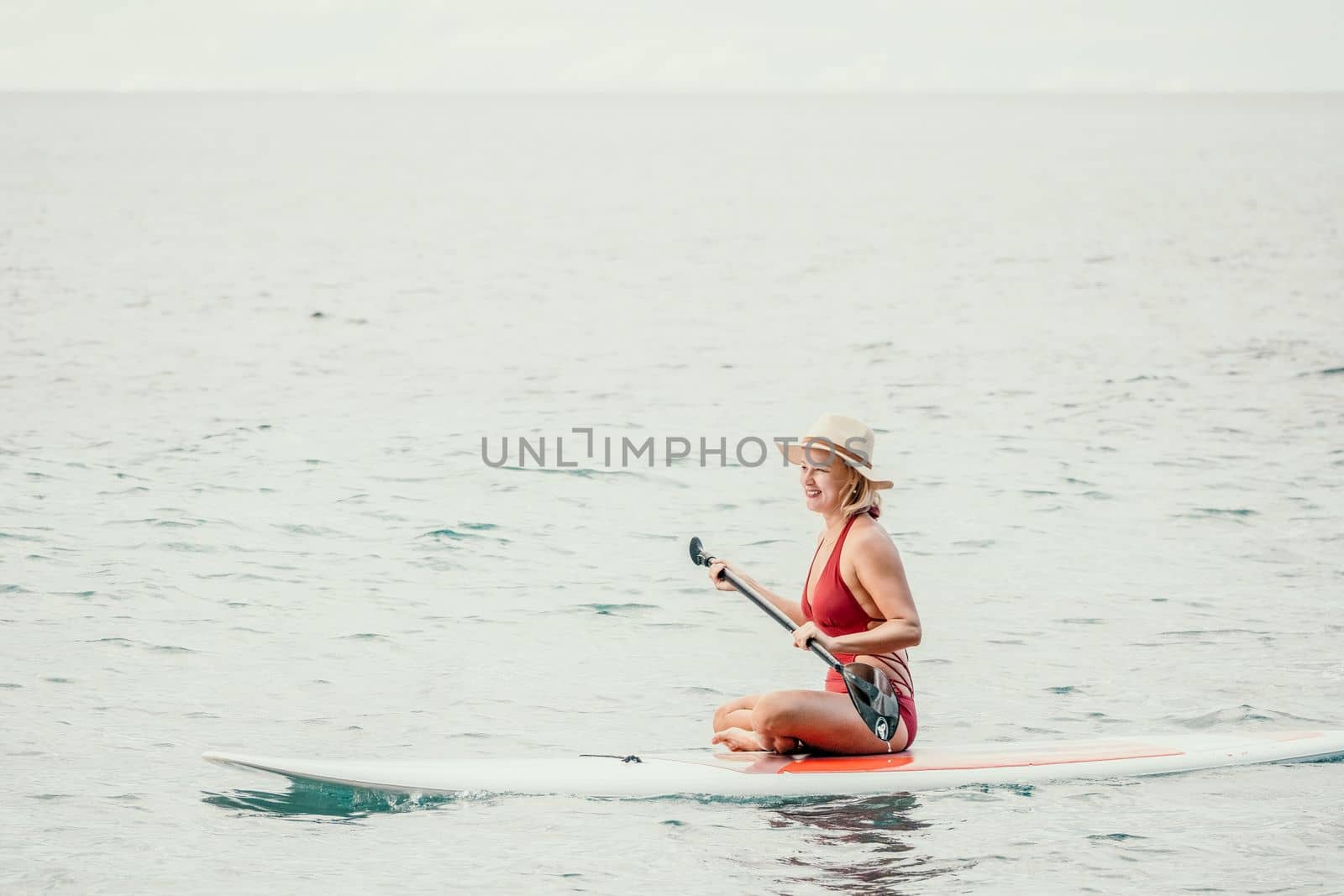Woman in red bikini on sup board. Happy lady with blond hair in red bathing suit chilling and sunbathing by turquoise sea ocean on sunset. Holiday, vacation and recreational concept. by panophotograph