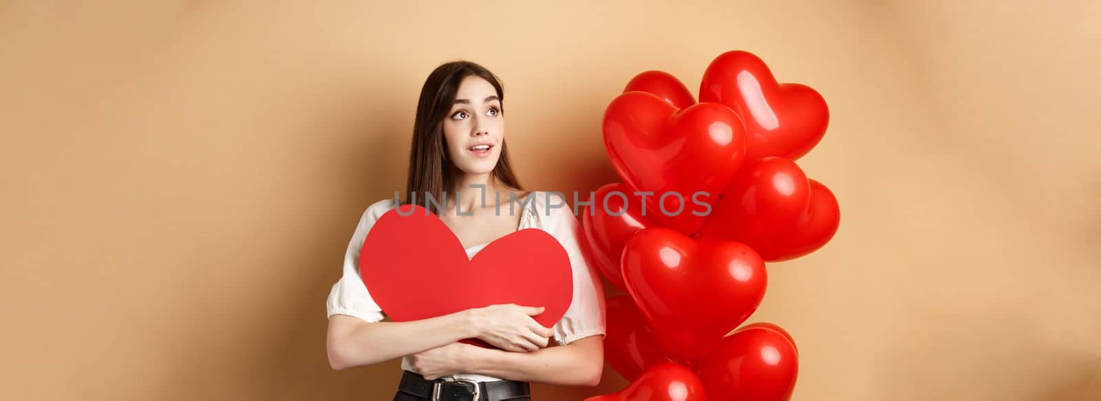 Romantic girl hugging Valentine day big red heart cutout, standing near romance balloons and looking left at logo, gazing dreamy at promo, beige background by Benzoix